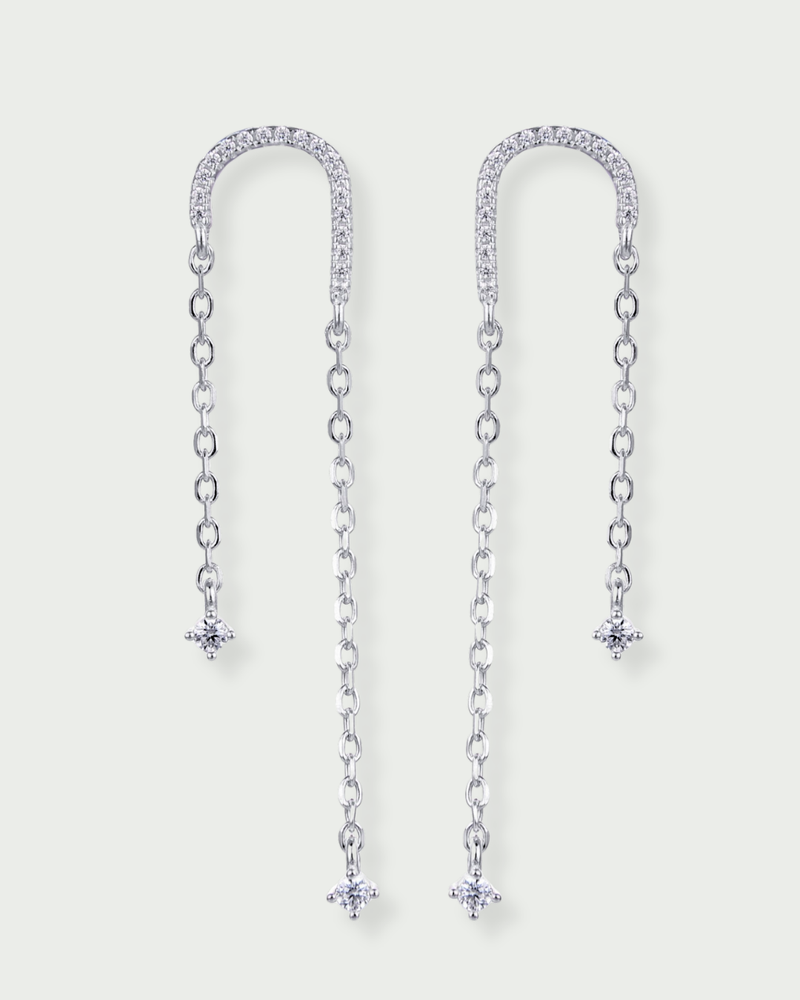 Boucles asymétriques en argent 925 rhodié, arc pavé de zirconiums et fines chaînes terminées par des pendants en pierres blanches.